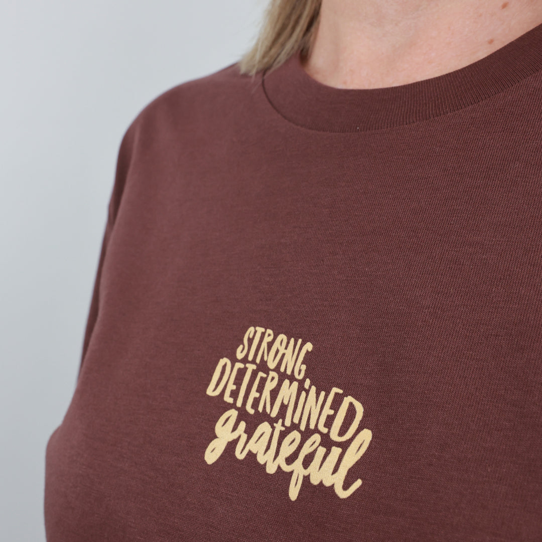 A close-up of a person wearing a brown crewneck sweatshirt with the words 'STRONG DETERMINED grateful' printed in a light beige script font on the chest.
