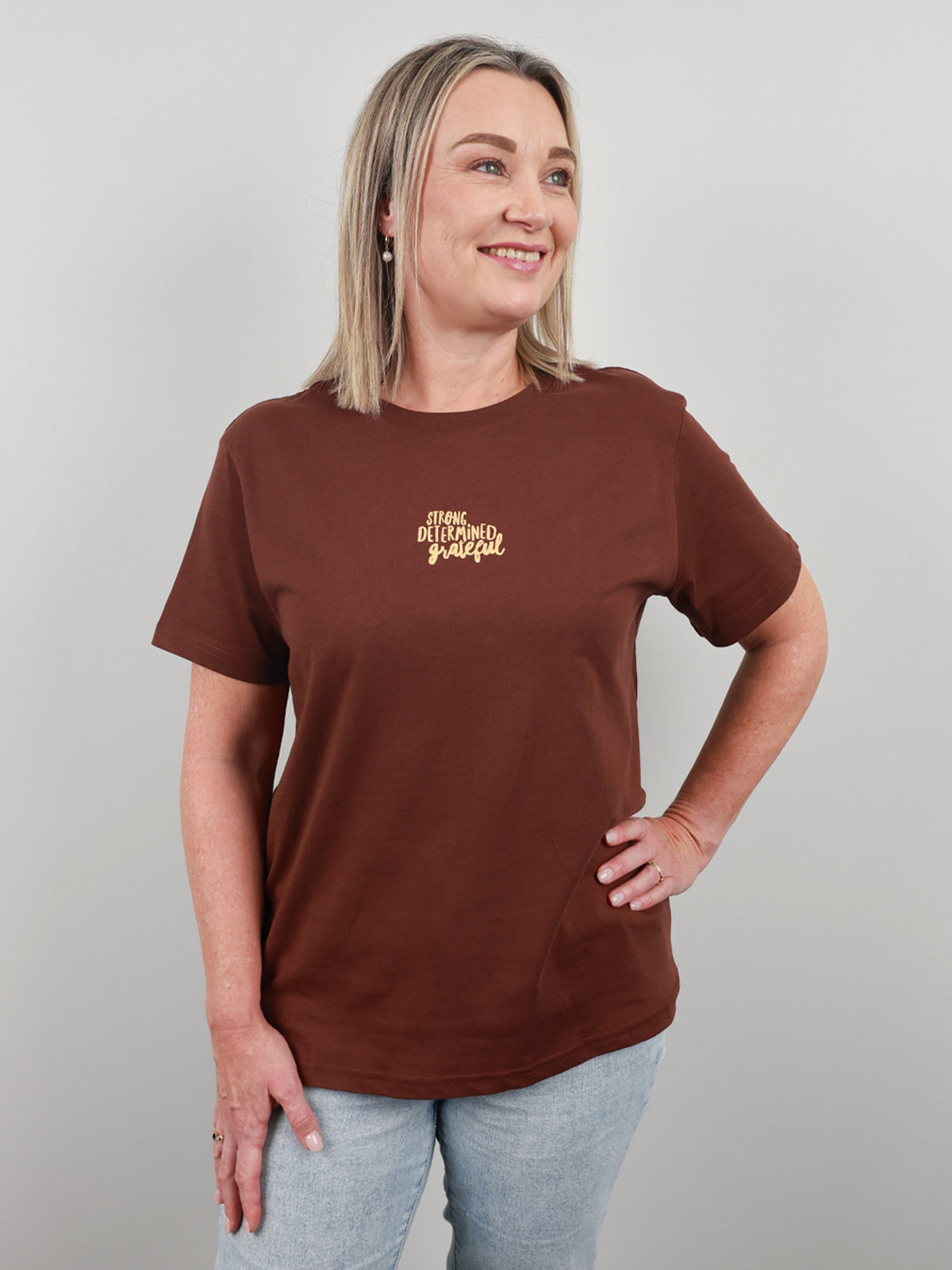 A woman wearing a brown short-sleeve t-shirt with a small gold graphic on the left chest, paired with light blue jeans, standing against a solid gray studio background.