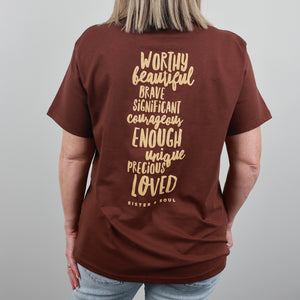 A woman shown from the back wearing a brown short-sleeve t-shirt with a gold typographic quote printed on the back, paired with light blue jeans against a neutral gray background.