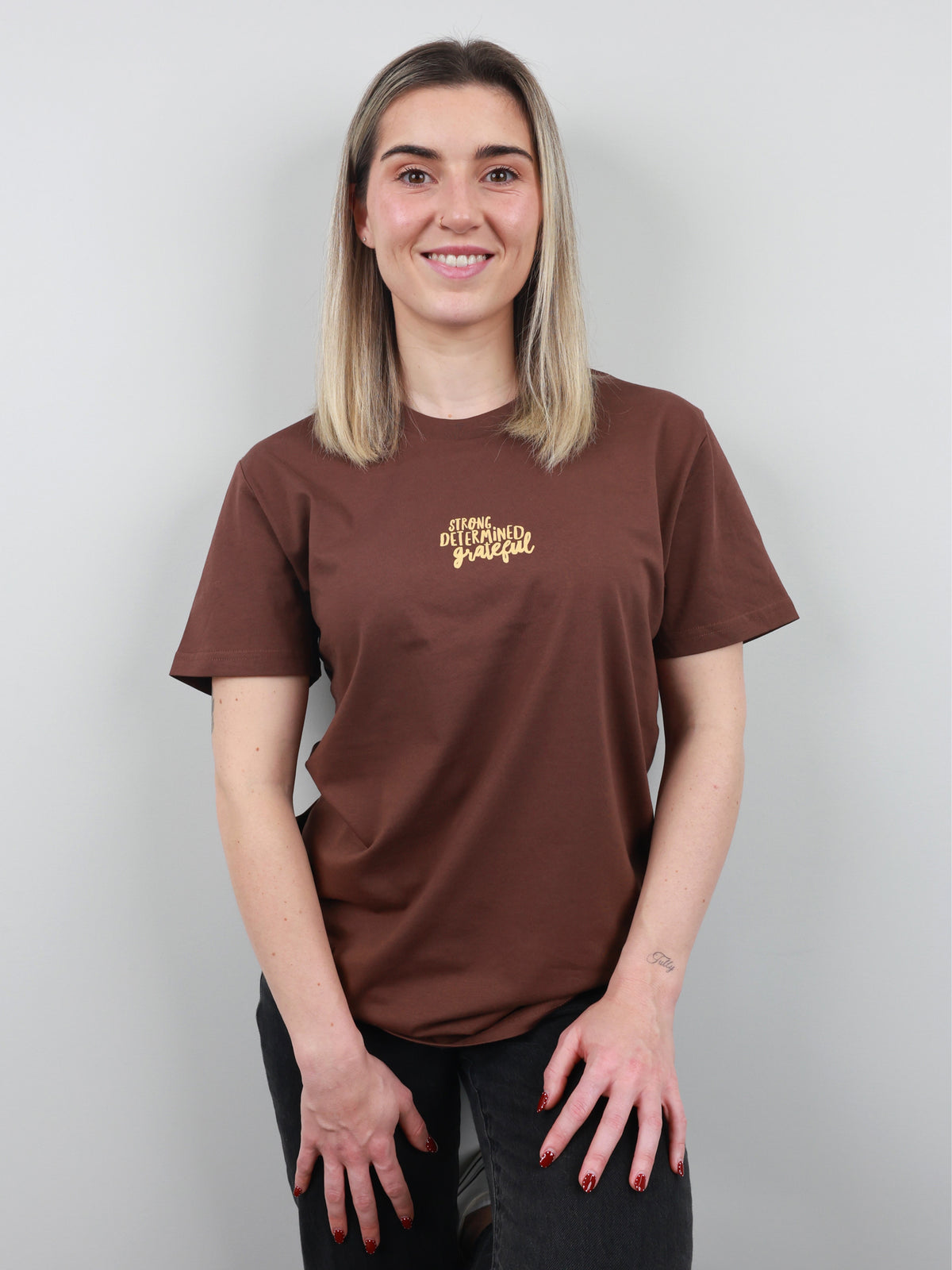 A woman with blonde hair wearing a brown t-shirt with a gold graphic on the left chest, standing against a light gray background.