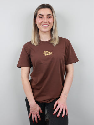 A woman with blonde hair wearing a brown t-shirt with a gold graphic on the left chest, standing against a light gray background.