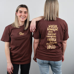 Two women modeling a brown short-sleeve graphic T-shirt against a light gray studio background. One shows the front with a small gold chest print, and the other shows the back with a large gold typographic quote.