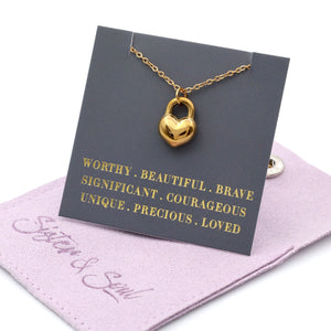 Gold heart-shaped lock necklace on a card with motivational phrases, displayed on a pink fabric background.