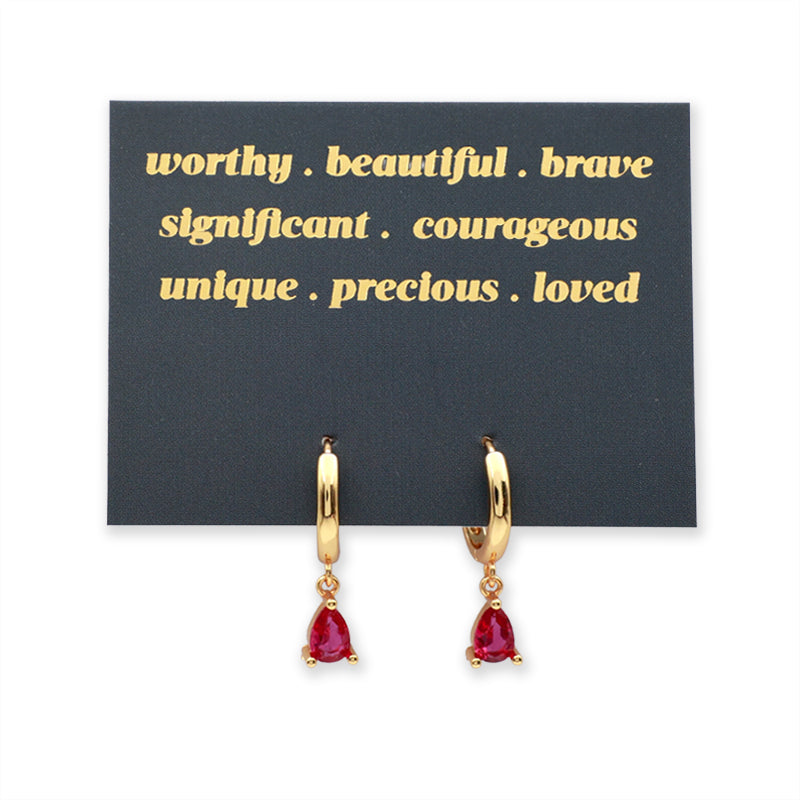 A pair of gold huggie hoop earrings with dangling dark pink teardrop gemstones, displayed on a black card with gold text that reads 'worthy. beautiful.