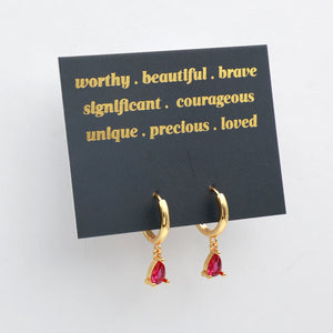 Gold huggie hoop earrings with dangling teardrop-shaped dark pink gemstones, displayed on a black card with gold text against a white background.