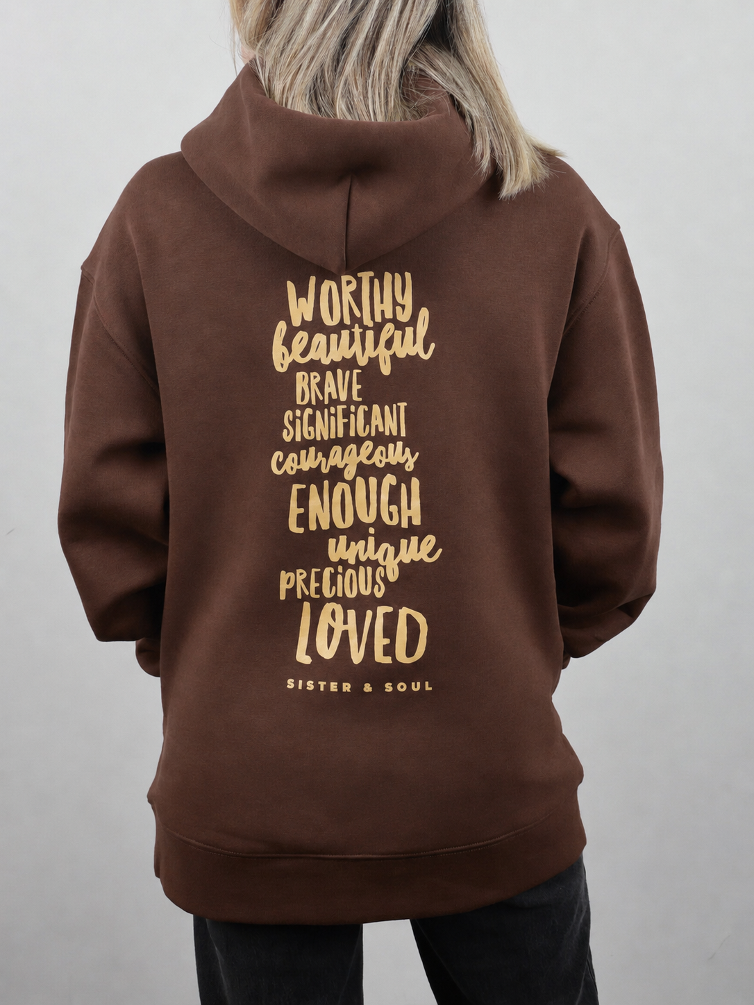 A person shown from the back wearing a brown hoodie with a gold typographic quote printed on the back. The text reads 'WORTHY beautiful BRAVE SIGNIFICANT courageously ENOUGH unique precious LOVED' with 'SISTER & SOUL' underneath.