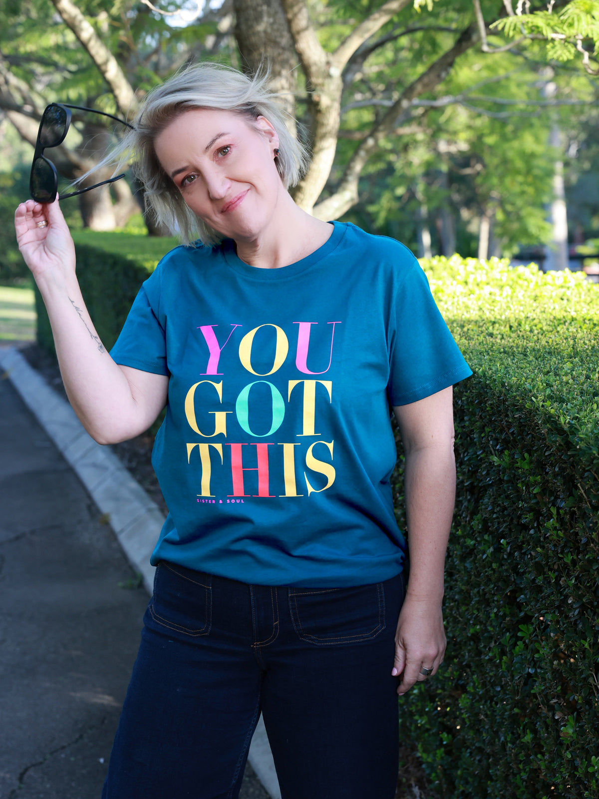 A woman wearing a teal graphic t-shirt with the text 'YOU GOT THIS' in colorful gradient letters, standing outdoors in front of a green hedge and trees.