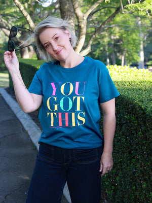 A woman wearing a teal graphic t-shirt with the text 'YOU GOT THIS' in colorful gradient letters, standing outdoors in front of a green hedge and trees.