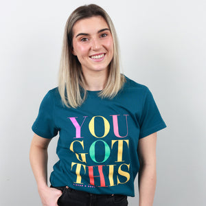 A smiling woman with blonde-highlighted hair wearing a teal t-shirt with the text 'YOU GOT THIS' printed in large, colorful gradient letters. The text features pink, yellow, green, and red colors.