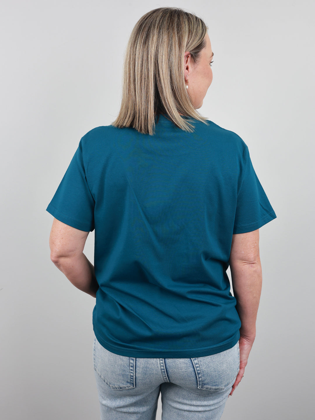 A woman with blonde hair is shown from the back wearing a teal blue short-sleeve t-shirt and light wash jeans against a plain light gray background.