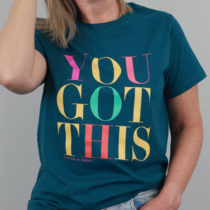 A woman wearing a dark teal graphic t-shirt with the text 'YOU GOT THIS' printed in large, colorful retro letters (pink, yellow, green, red) on the front. The brand 'SISTER & SOUL' is printed in small letters below the text.