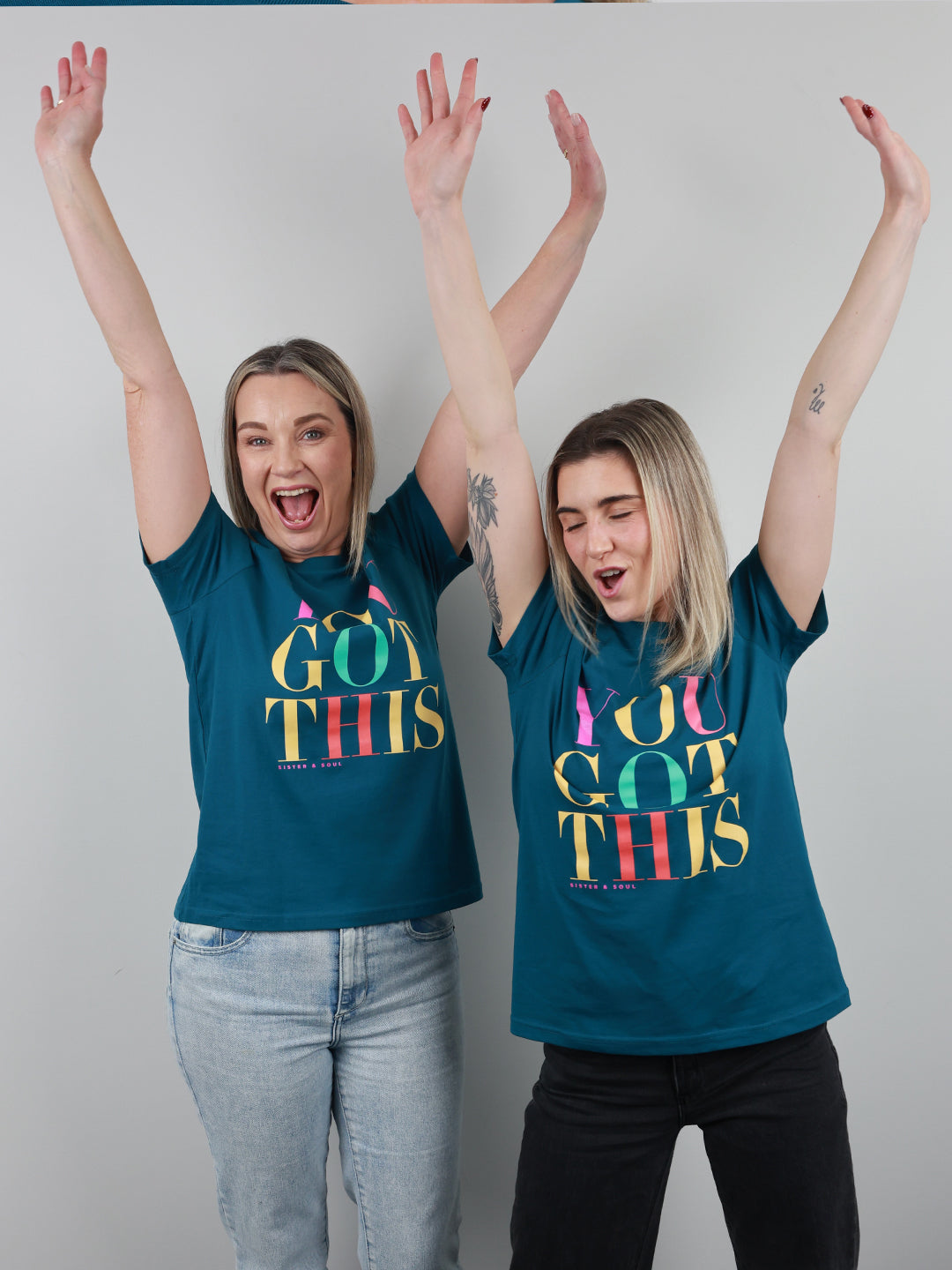 Two women with raised arms wearing matching teal t-shirts featuring a colorful 'YOU GOT THIS' graphic, standing against a plain light gray background.