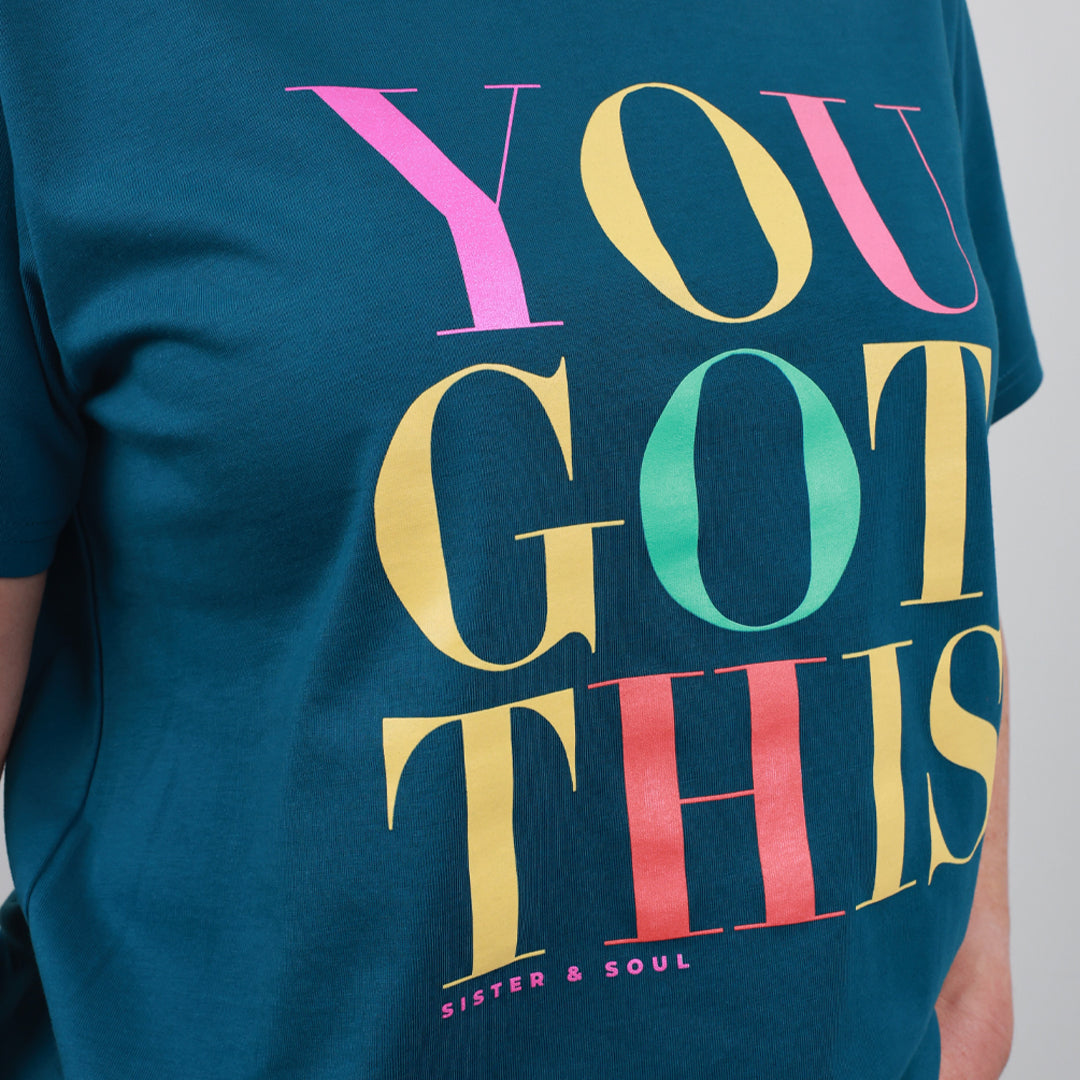 A close-up of a teal blue t-shirt featuring a large, colorful graphic print that reads 'YOU GOT THIS' in pink, yellow, green, and red letters, with 'SISTER & SOUL' printed below in small white text.