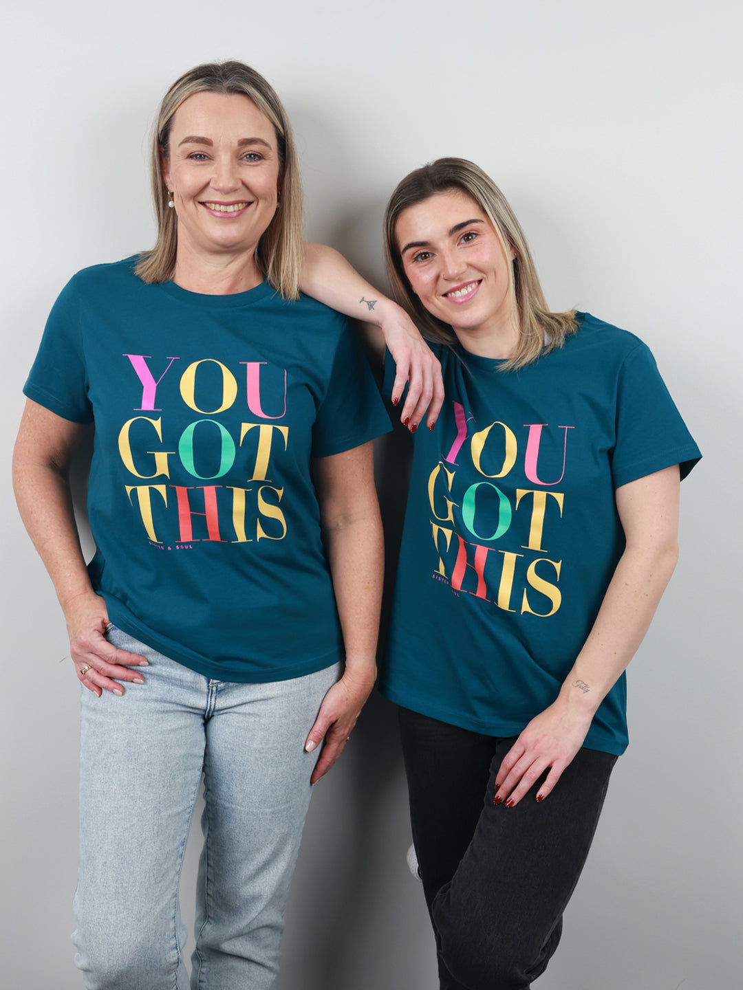 Two women standing side by side wearing matching teal t-shirts with a colorful 'YOU GOT THIS' graphic print, posed against a plain light gray wall.