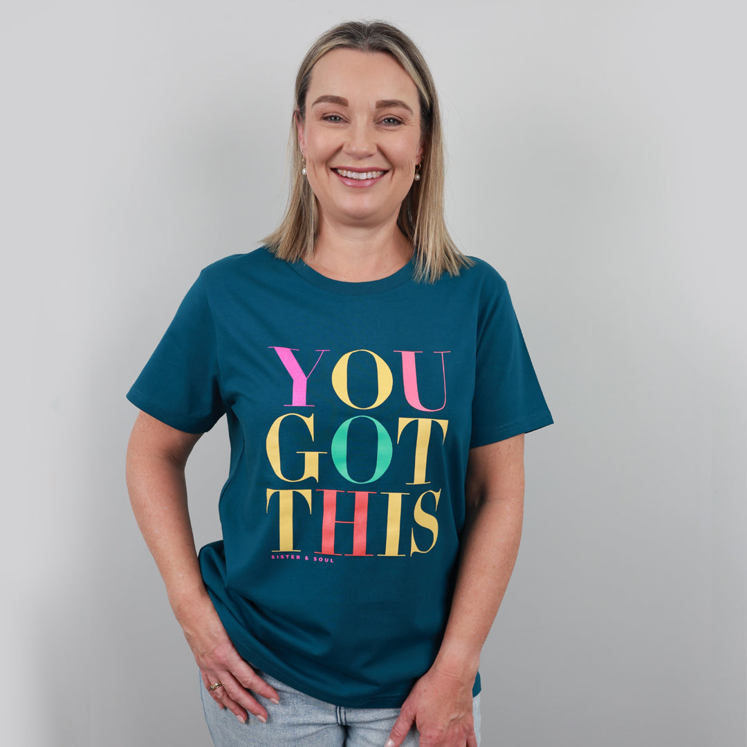 A smiling woman with blonde hair wearing a teal blue graphic t-shirt that reads 'YOU GOT THIS' in colorful block letters, paired with light wash jeans, standing against a neutral gray background.