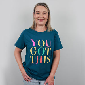 A smiling woman with blonde hair wearing a teal blue graphic t-shirt that reads 'YOU GOT THIS' in colorful block letters, paired with light wash jeans, standing against a neutral gray background.