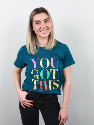 A smiling woman with blonde hair wearing a teal graphic t-shirt that reads 'YOU GOT THIS' in colorful letters, paired with black jeans, standing against a light gray background.