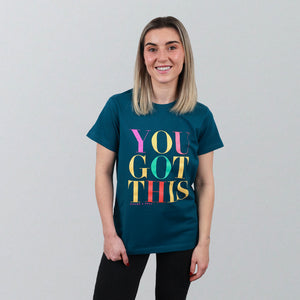 A woman with blonde hair wearing a dark teal graphic t-shirt that reads 'YOU GOT THIS' in colorful letters, standing against a light gray background.