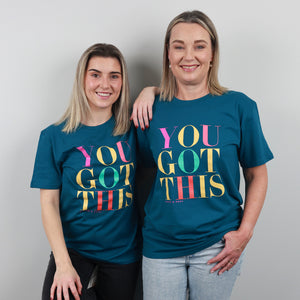 Two women standing side by side wearing matching teal graphic t-shirts with the text 'YOU GOT THIS' in colorful letters. The woman on the right is wearing a larger size and the woman on the left is wearing a smaller size of the same shirt.