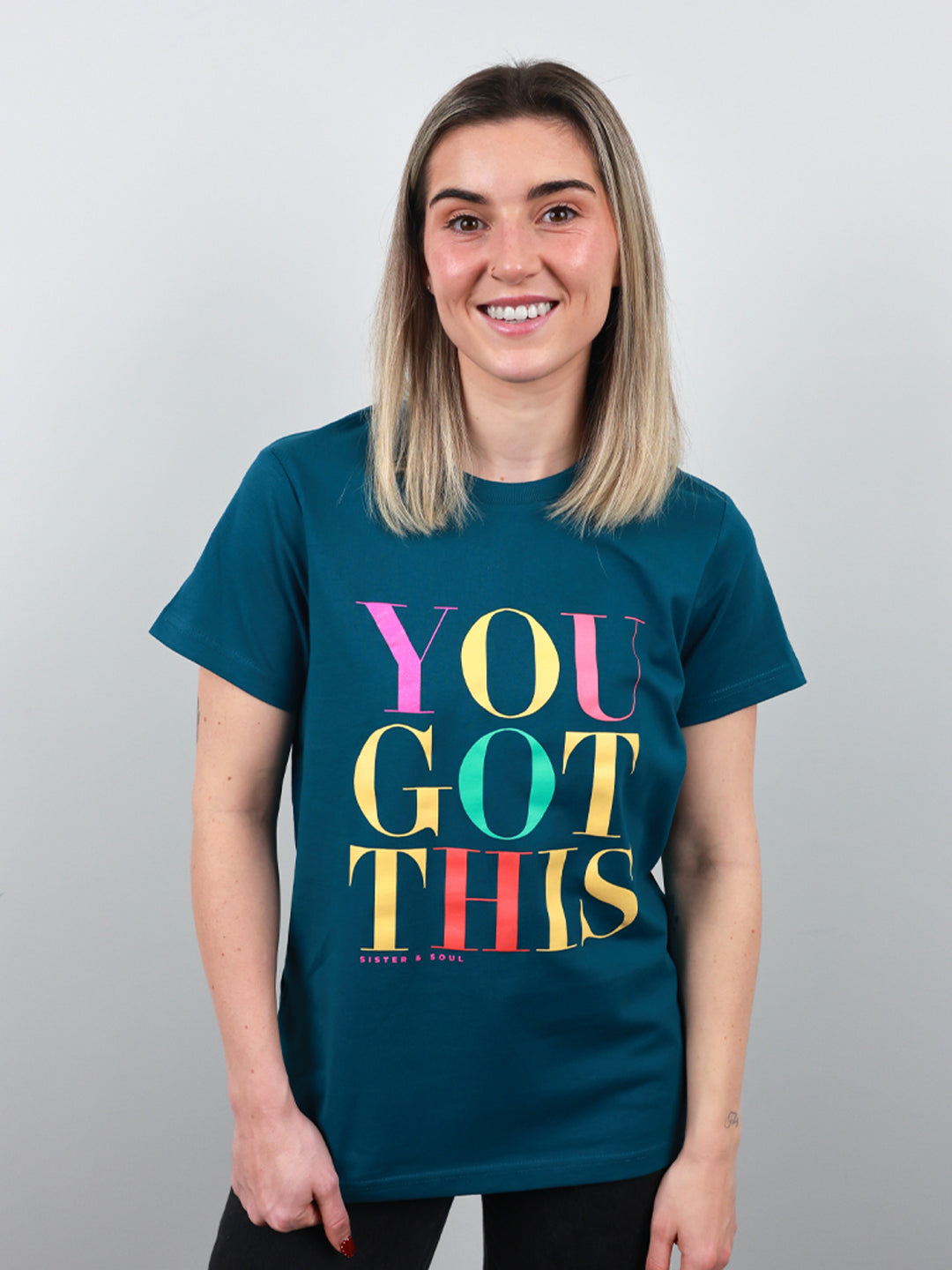 A smiling woman with blonde hair wearing a dark teal t-shirt with the text 'YOU GOT THIS' printed in colorful retro letters, set against a plain light gray background.