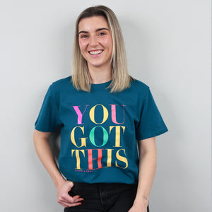A smiling woman with shoulder-length blonde hair wears a teal graphic t-shirt featuring the text 'YOU GOT THIS' in large, colorful retro letters. She is standing against a plain light gray wall.
