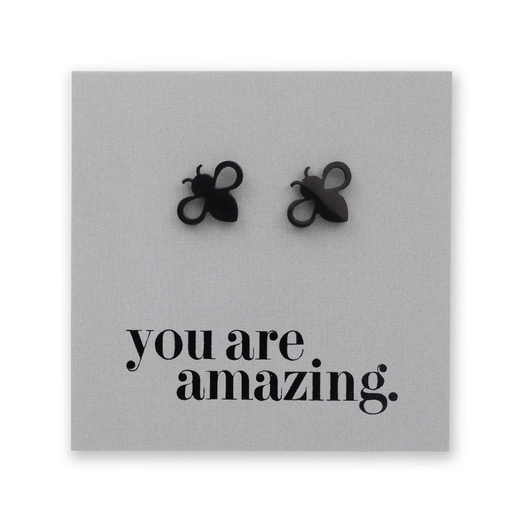 A pair of black bee-shaped stud earrings displayed on a square gray backing card that reads 'you are amazing.' in black text.