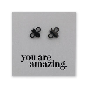 A pair of black bee-shaped stud earrings displayed on a square gray backing card that reads 'you are amazing.' in black text.