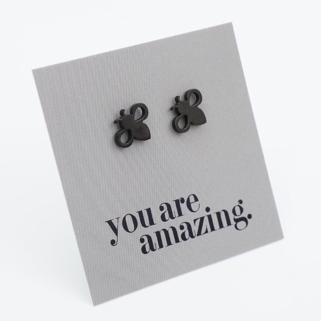 A pair of small black bee-shaped stud earrings displayed on a textured gray square card that reads 'you are amazing.' in black script, photographed on a clean white background.