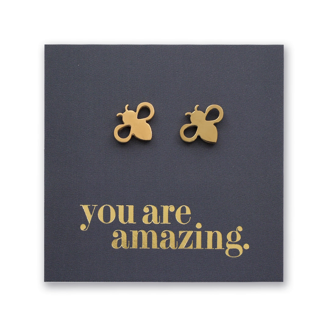 A pair of small gold bee stud earrings displayed on a dark charcoal square card with the phrase 'you are amazing.' in gold lettering, shown against a white background.