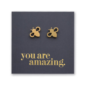 A pair of small gold bee stud earrings displayed on a dark charcoal square card with the phrase 'you are amazing.' in gold lettering, shown against a white background.
