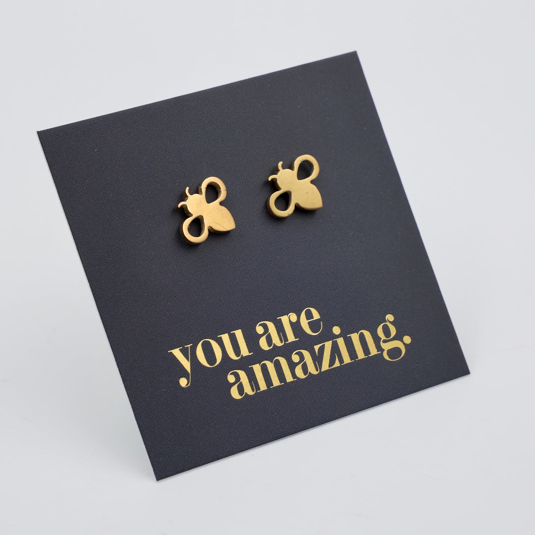 Four pairs of small bee-shaped stud earrings displayed on dark gray cards with the phrase 'you are amazing.' in different metallic colors.