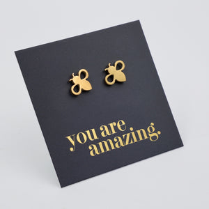 A pair of small gold bee stud earrings displayed on a grey  backing card with the phrase 'you are amazing.' in gold lettering, photographed on a white  background.