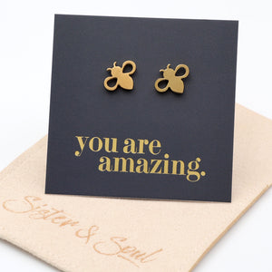 A pair of gold bee stud earrings displayed on a black backing card that reads 'you are amazing.' in gold script. on a oat / beige coloured pouch 