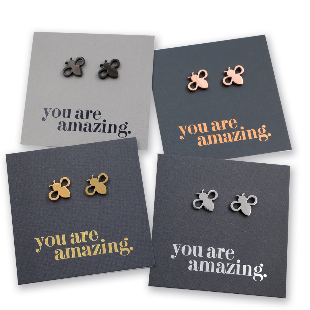 Four pairs of small bee-shaped stud earrings displayed on dark gray cards with the phrase 'you are amazing.' in different metallic colors.