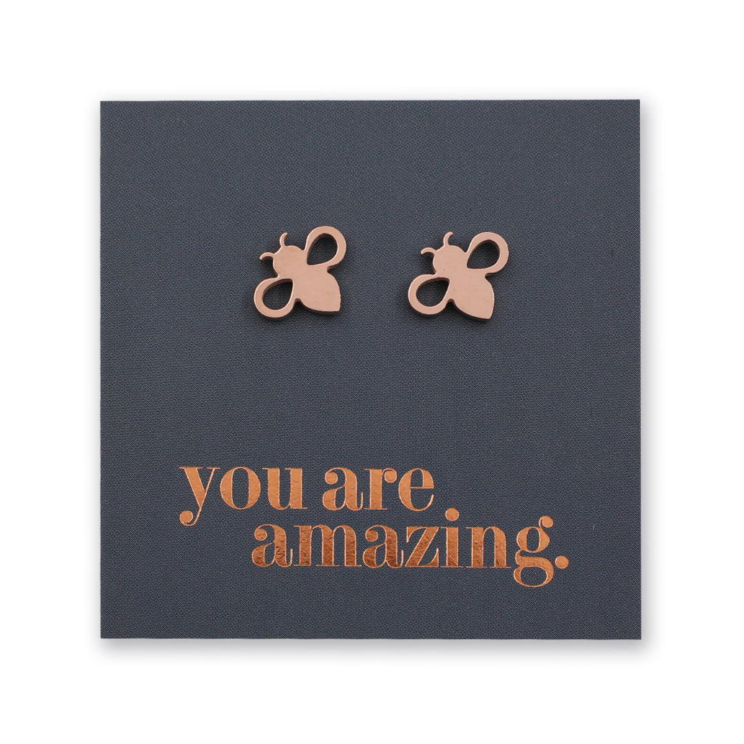 A pair of small bee-shaped stud earrings in a metallic rose gold tone, displayed on a dark charcoal square card with the phrase 'you are amazing.' in matching metallic lettering.