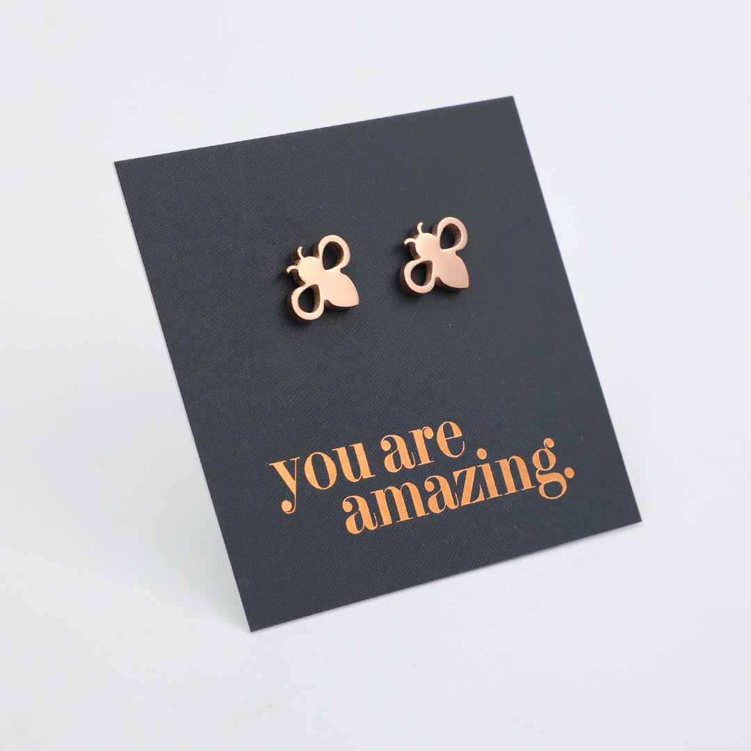 Rose gold bee stud earrings displayed on a grey backing card with the words 'you are amazing.' in rose gold lettering, photographed on a light white background.