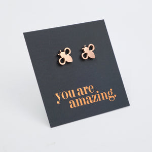 Rose gold bee stud earrings displayed on a grey backing card with the words 'you are amazing.' in rose gold lettering, photographed on a light white background.