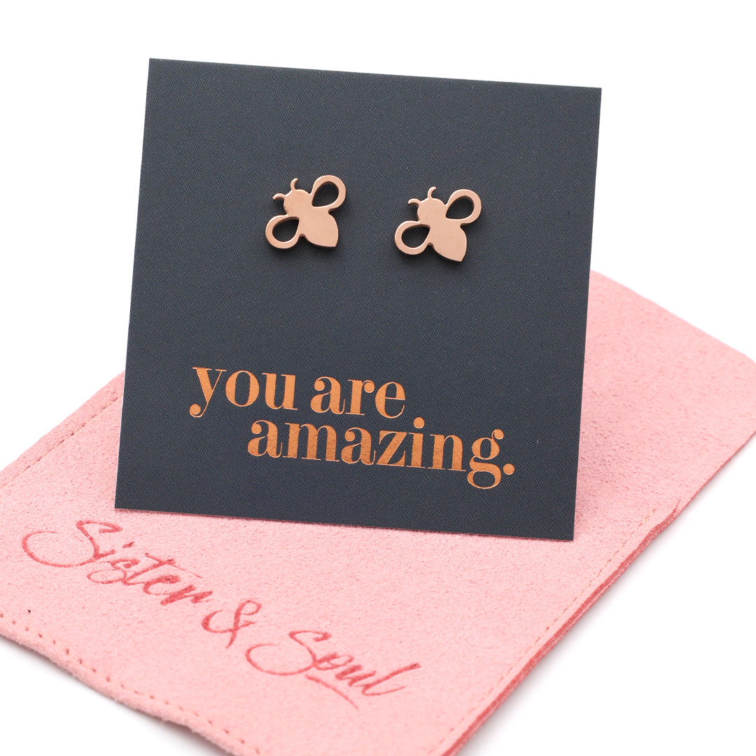 Rose gold bee stud earrings displayed on a black card that reads 'you are amazing.' with a pink fabric pouch underneath on a white background.