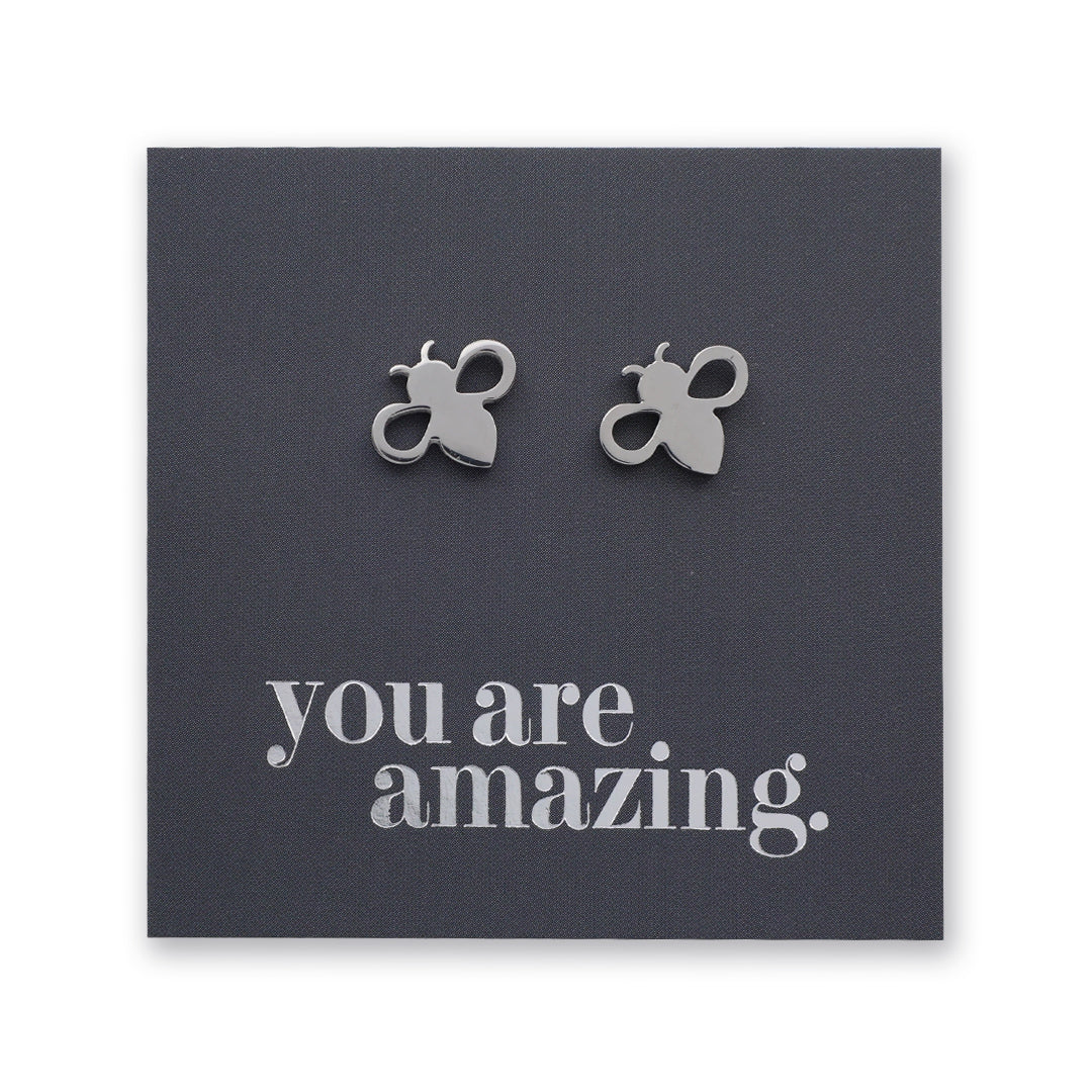 A pair of small silver bee stud earrings displayed on a dark gray square backing card that reads 'you are amazing.' in silver lowercase text.