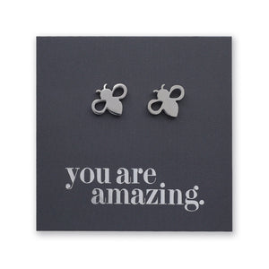 A pair of small silver bee stud earrings displayed on a dark gray square backing card that reads 'you are amazing.' in silver lowercase text.
