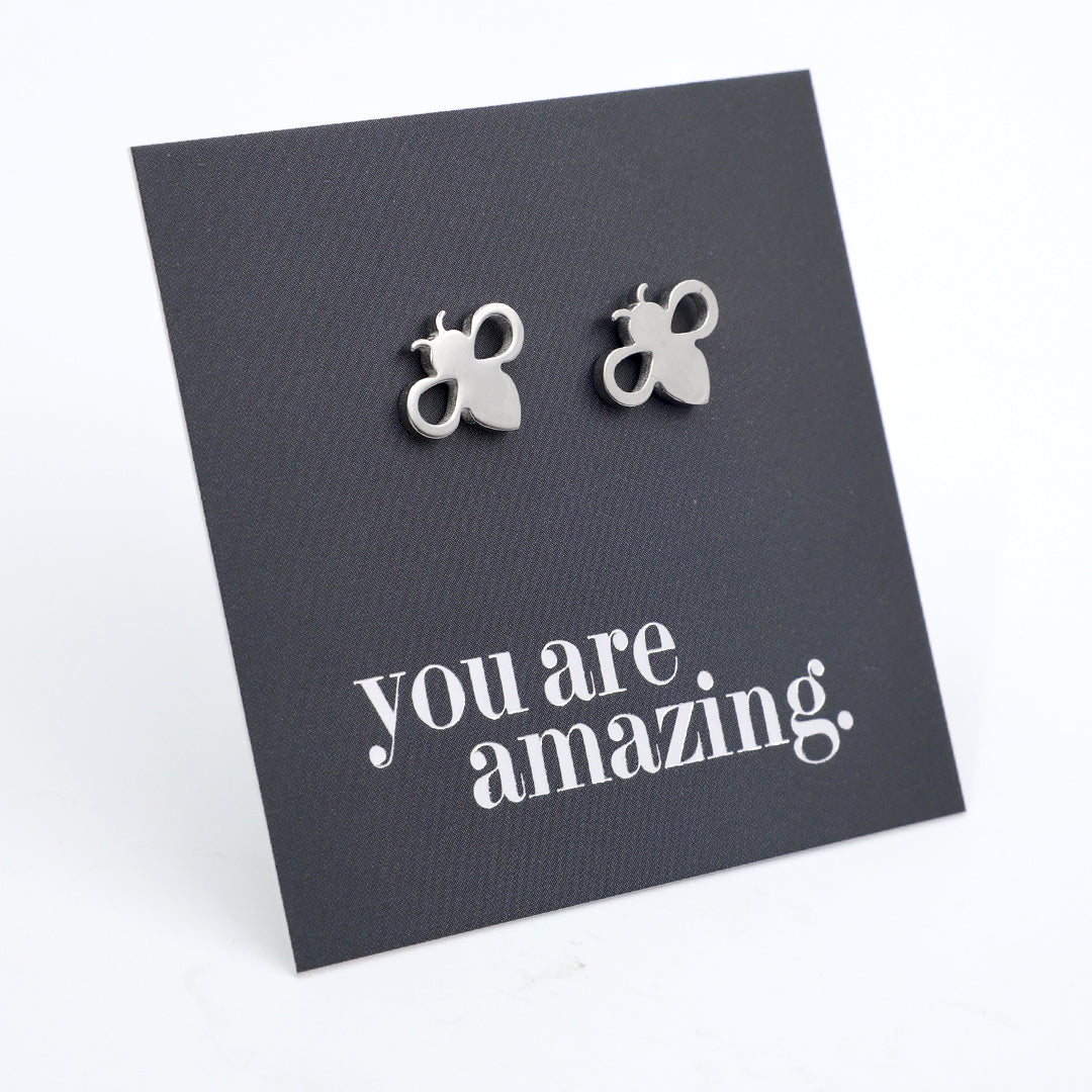 A pair of small silver bee stud earrings displayed on a dark gray square card that reads 'you are amazing.' in silver script, photographed on a clean white background.