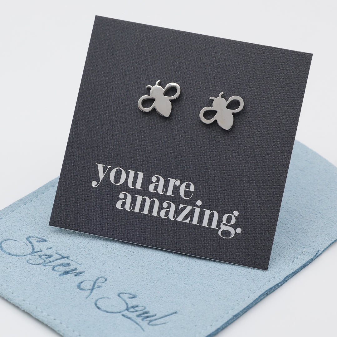 A pair of silver bee stud earrings displayed on a black backing card that reads 'you are amazing.' with a light blue fabric pouch underneath.