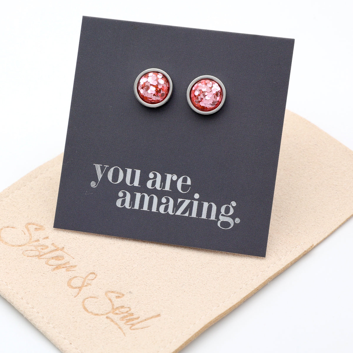 Pink glittery stud earrings on a card with 'you are amazing' text, displayed on a beige card with 'Sister & Swag' branding.