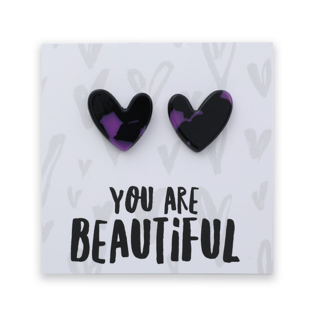 A pair of heart-shaped stud earrings with a glossy black and purple marbled pattern, mounted on a white display card that reads 'YOU ARE BEAUTIFUL' in black text.