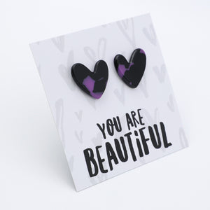 A pair of heart-shaped stud earrings with a black and purple marbled pattern, displayed on a white card that reads 'YOU ARE BEAUTIFUL' in black lettering.