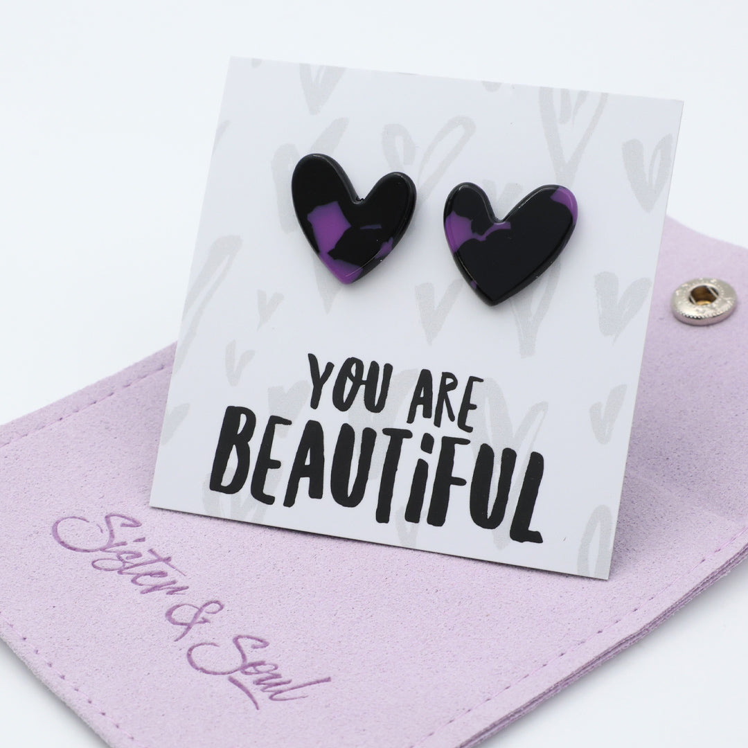 A pair of heart-shaped stud earrings with a black and purple marbled pattern, displayed on a white backing card that reads 'YOU ARE BEAUTIFUL'. The card is resting on a light purple fabric pouch with the brand name 'Sister & Soul' embroidered on it.
