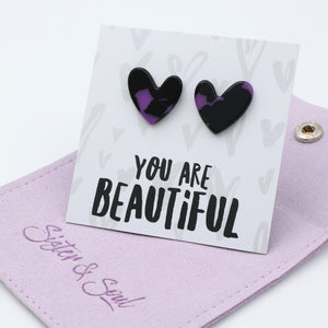 A pair of heart-shaped stud earrings with a black and purple marbled pattern, displayed on a white backing card that reads 'YOU ARE BEAUTIFUL'. The card is resting on a light purple fabric pouch with the brand name 'Sister & Soul' embroidered on it.