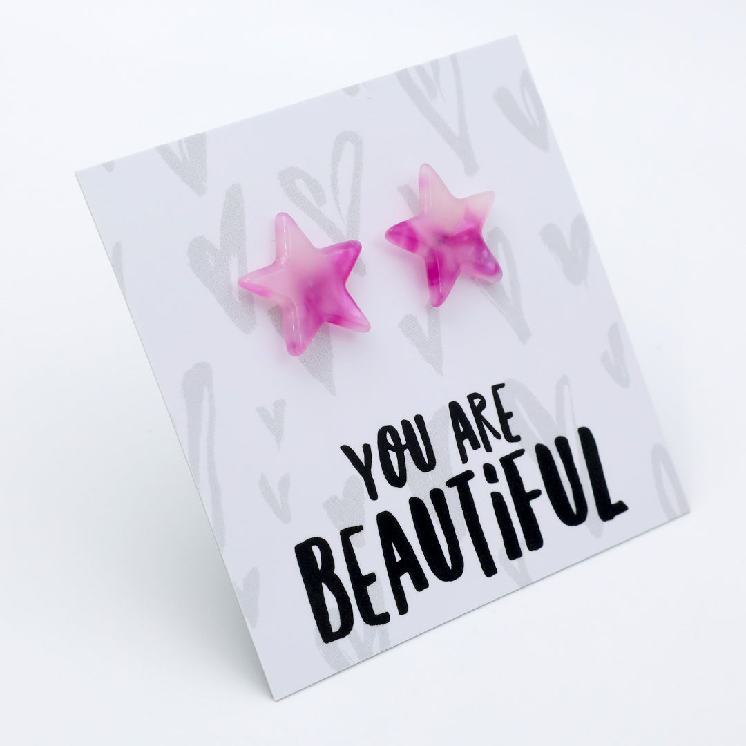 A pair of pink star-shaped stud earrings displayed on a white card that reads 'YOU ARE BEAUTIFUL' in black lettering, photographed on a clean white background.