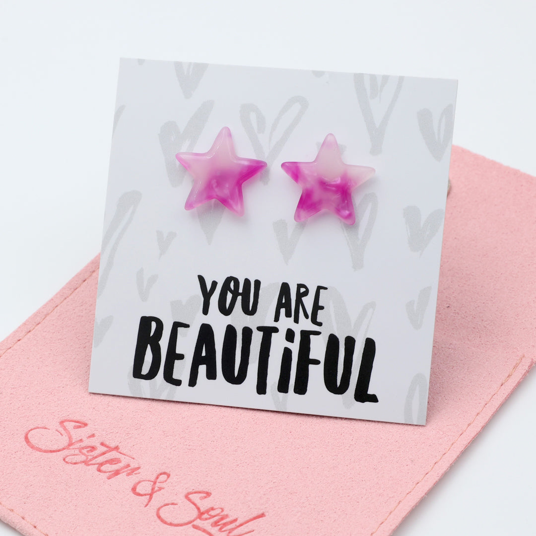 Two pink marbled star-shaped stud earrings displayed on a white card that reads 'YOU ARE BEAUTIFUL', resting on a pink fabric pouch with ' Sister & Soul' in pink.