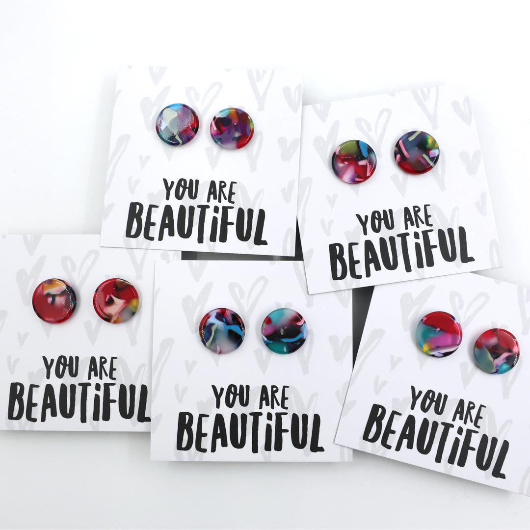 A pair of round acrylic stud earrings with a colorful marbled pattern in red, pink, blue, and yellow, displayed on a white backing card that reads 'YOU ARE BEAUTIFUL' in black text.